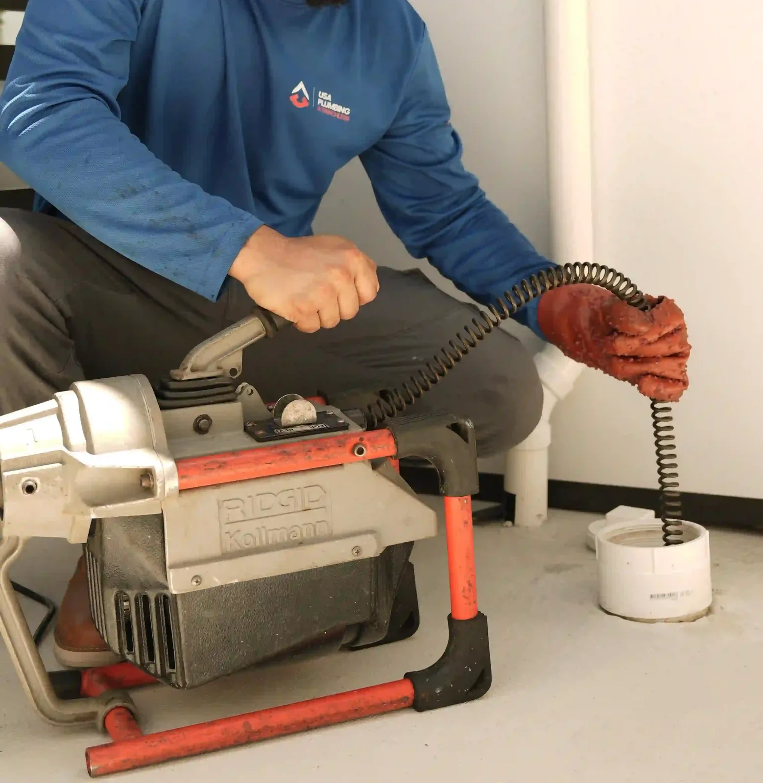 A plumber crouched over a RIDGID drum machine, feeding the coiled steel cable by red-gloved hand into an open white PVC floor cleanout beside an interior wall, with the drum ready to spin a cutting head down the line